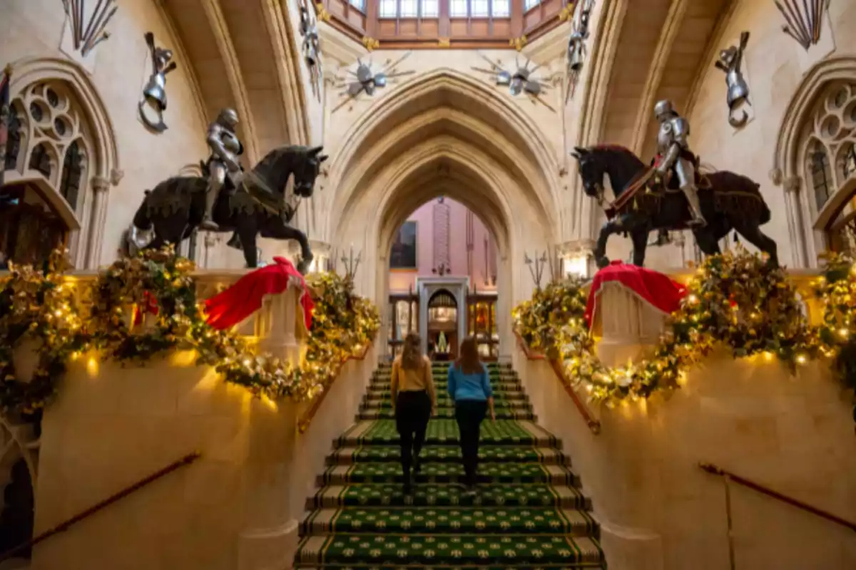 Dos personas suben una escalera decorada con guirnaldas y luces navideñas en un edificio con arquitectura gótica y estatuas de caballeros en armadura montados en caballos.