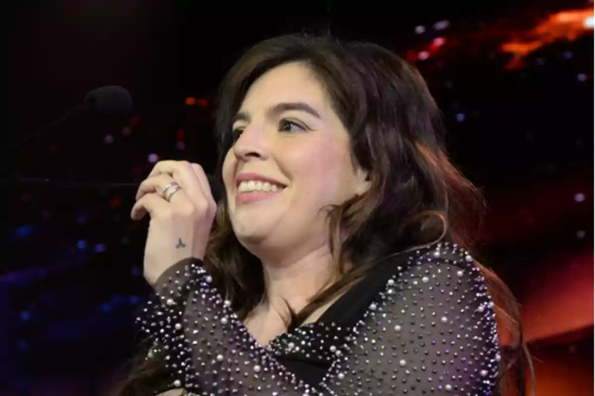 Mujer sonriendo mientras sostiene un micrófono en un escenario iluminado. Mujer sonriendo mientras sostiene un micrófono en un escenario iluminado.