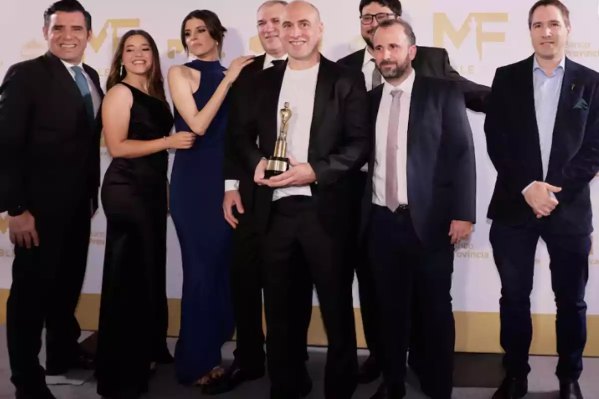 Un grupo de personas elegantemente vestidas posando en un evento, una de ellas sostiene un trofeo. Un grupo de personas elegantemente vestidas posando en un evento, una de ellas sostiene un trofeo.