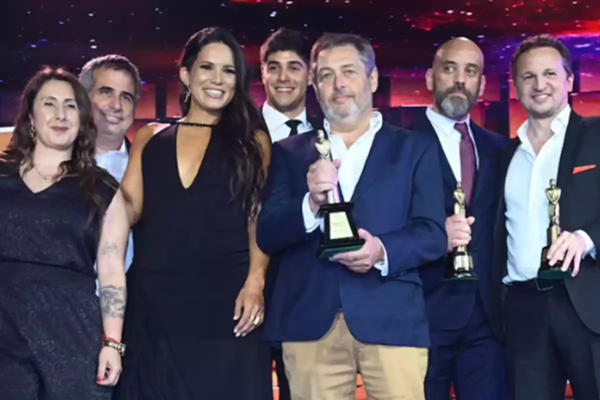 Un grupo de personas sonríe mientras sostiene trofeos en un evento con un fondo iluminado. Un grupo de personas sonríe mientras sostiene trofeos en un evento con un fondo iluminado.