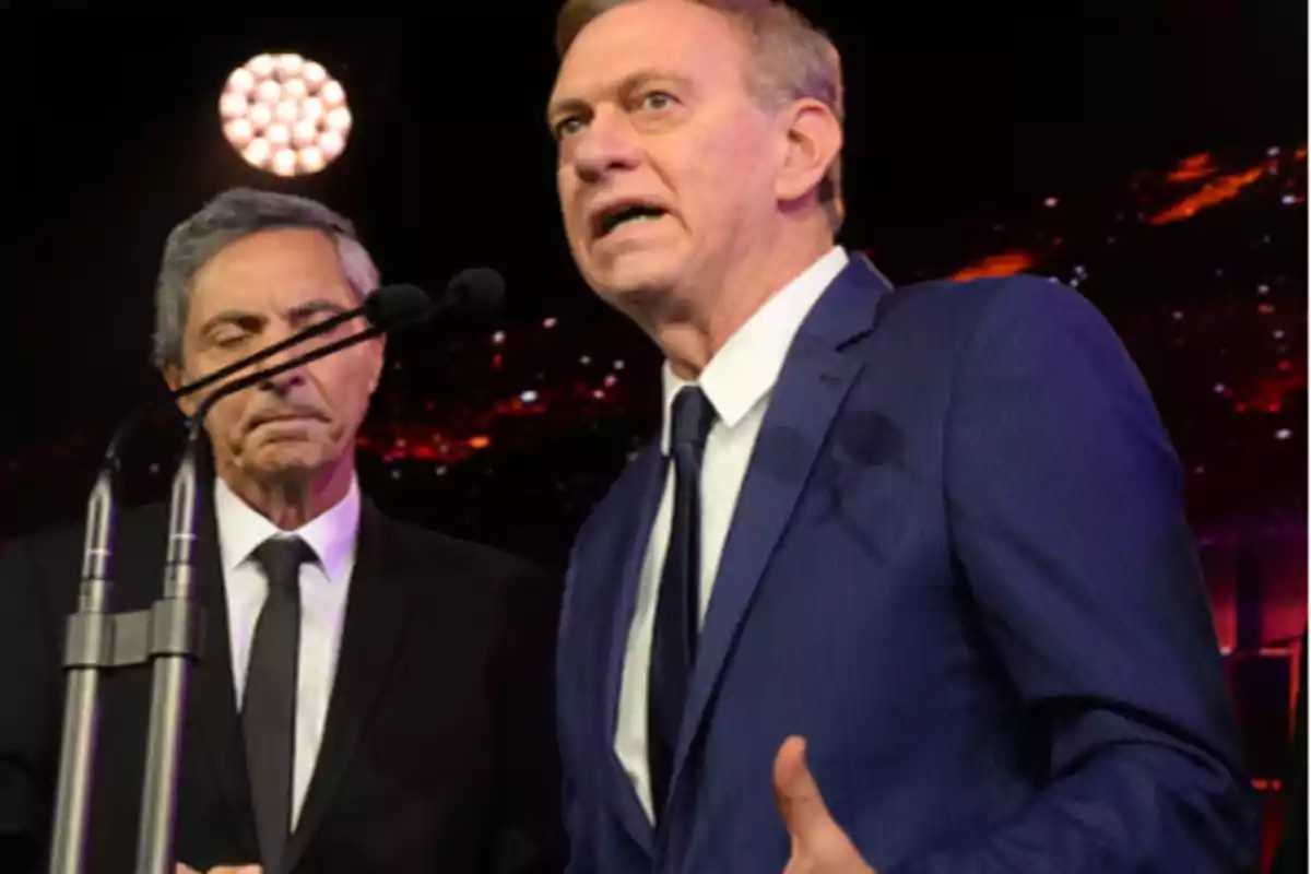 Dos hombres en trajes formales hablando en un escenario con micrófonos y luces de fondo. Dos hombres en trajes formales hablando en un escenario con micrófonos y luces de fondo.