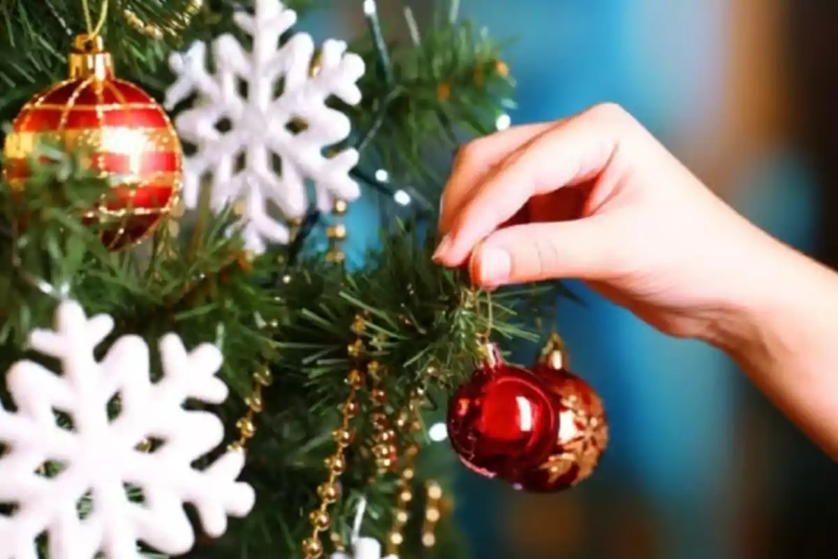 Una mano colocando una esfera roja en un árbol de Navidad decorado con copos de nieve blancos y guirnaldas doradas.