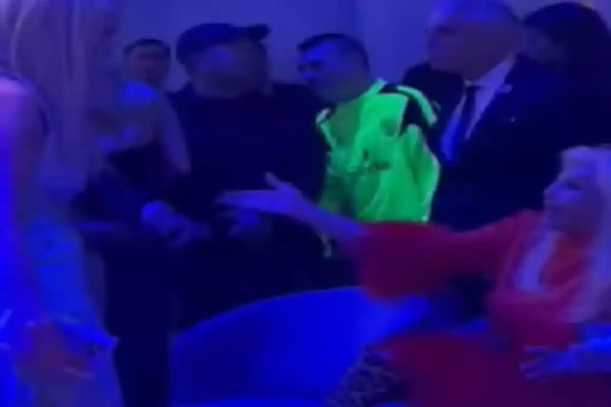 Un grupo de personas en un ambiente iluminado con luces azules, una persona vestida de rojo está sentada y parece estar extendiendo la mano hacia otra persona de pie. Un grupo de personas en un ambiente iluminado con luces azules, una persona vestida de rojo está sentada y parece estar extendiendo la mano hacia otra persona de pie.