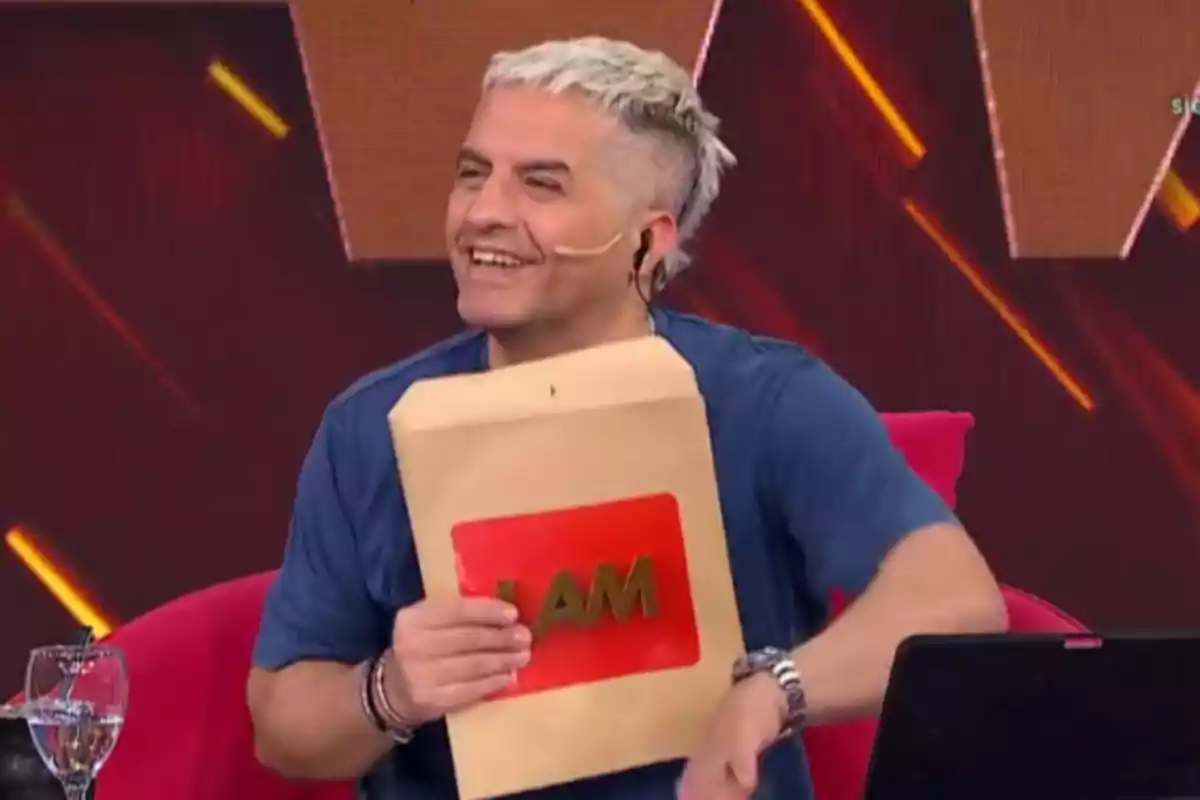 Un presentador de televisión sonríe mientras sostiene un sobre con el logo de LAM en un set de grabación.