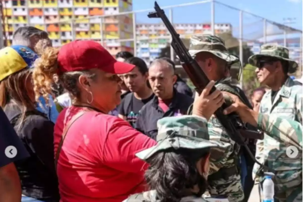 Un grupo de personas, algunas con uniformes militares, interactúa en un espacio al aire libre con edificios coloridos al fondo.