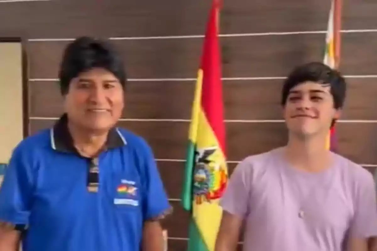 Dos personas posan frente a una bandera de Bolivia en un interior con paredes de madera.