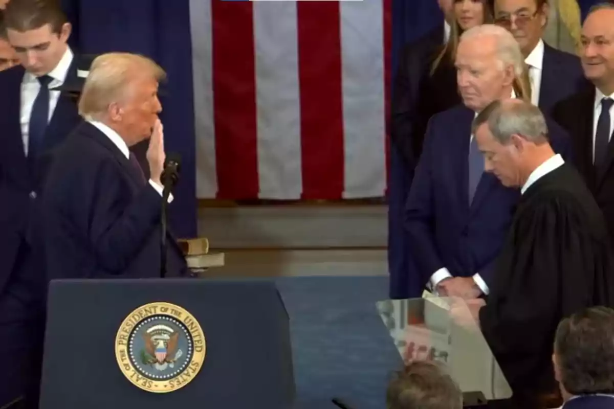 Un grupo de personas en un evento formal con banderas de fondo, donde un hombre está realizando un juramento frente a un podio con el sello presidencial.