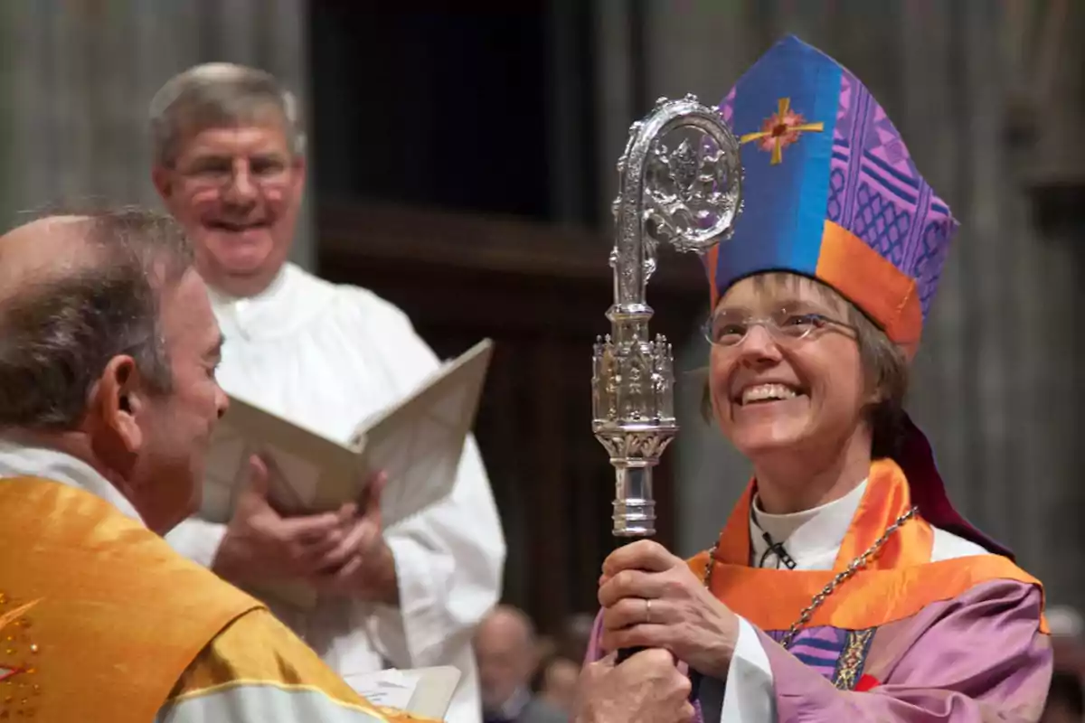 Una persona vestida con atuendo religioso colorido sostiene un báculo ceremonial mientras sonríe, acompañada de otras personas en un entorno eclesiástico.