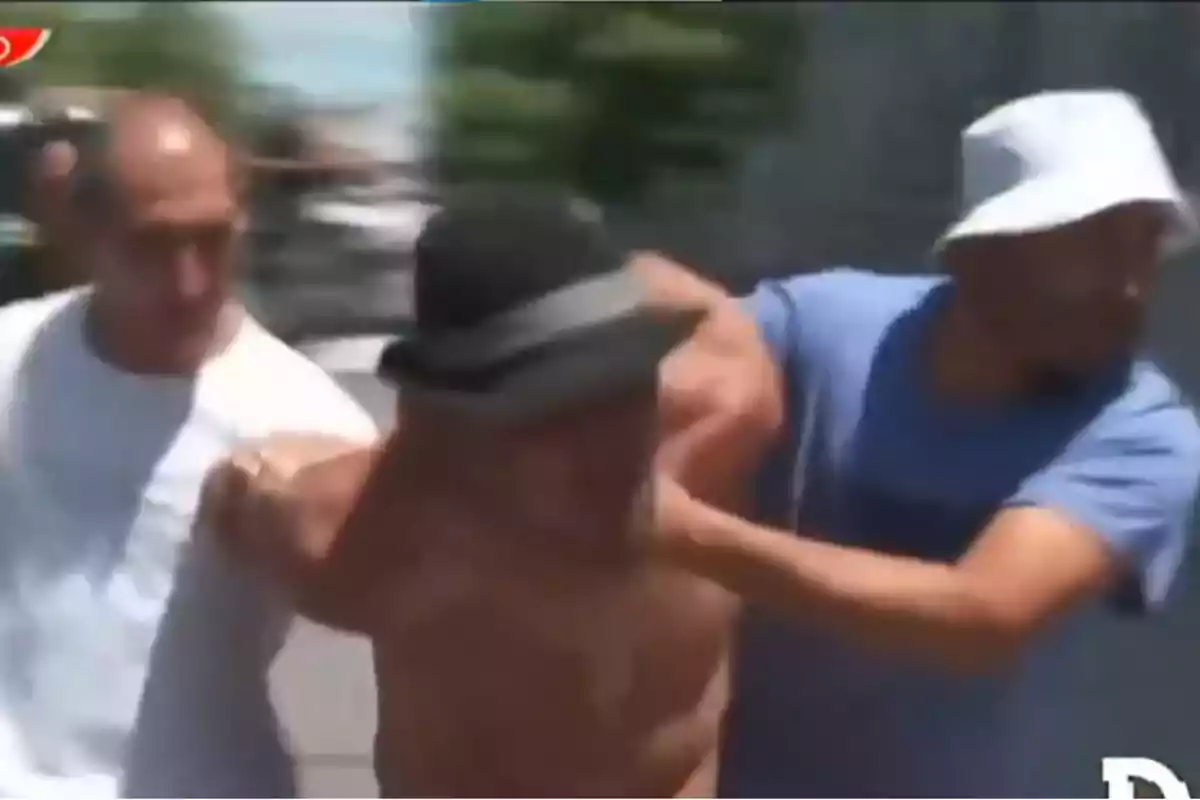 Tres personas en movimiento, una de ellas sin camisa y con sombrero, siendo sujetada por las otras dos.