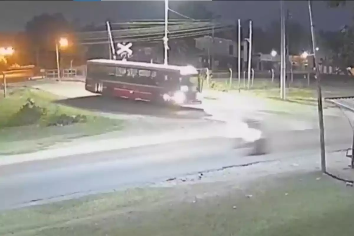 Un autobús cruza un paso a nivel durante la noche, iluminado por luces de la calle, mientras un vehículo se aproxima rápidamente.