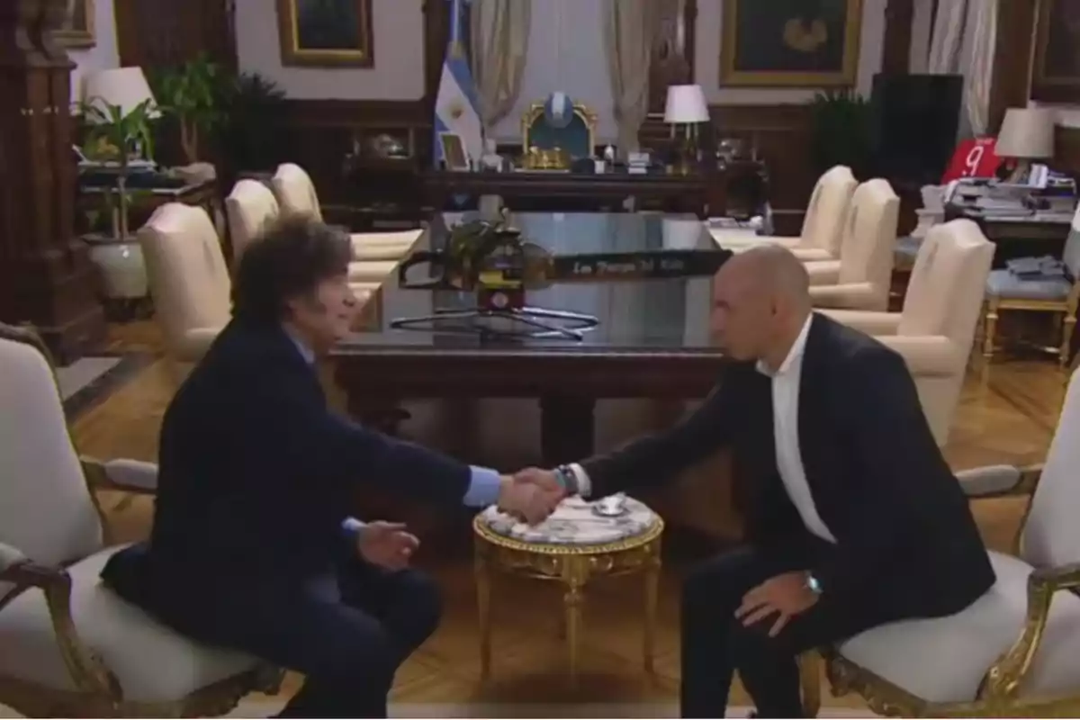 Dos personas se dan la mano en una sala elegante con muebles clásicos y una bandera de Argentina al fondo.