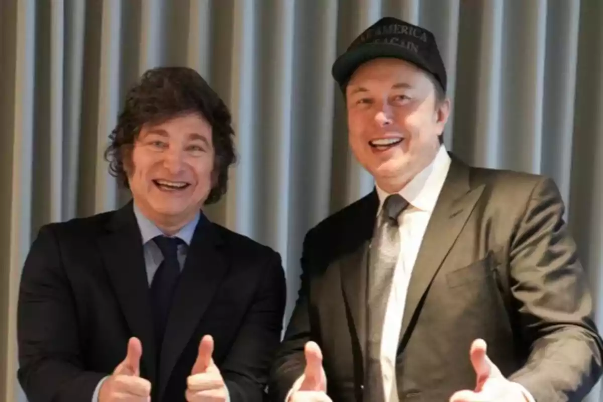 Dos hombres sonrientes con trajes oscuros levantando los pulgares frente a una cortina gris.