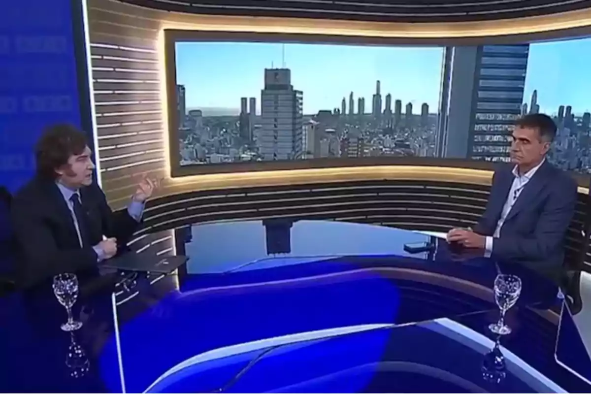 Dos personas sentadas en un estudio de televisión con una gran pantalla mostrando un paisaje urbano al fondo.