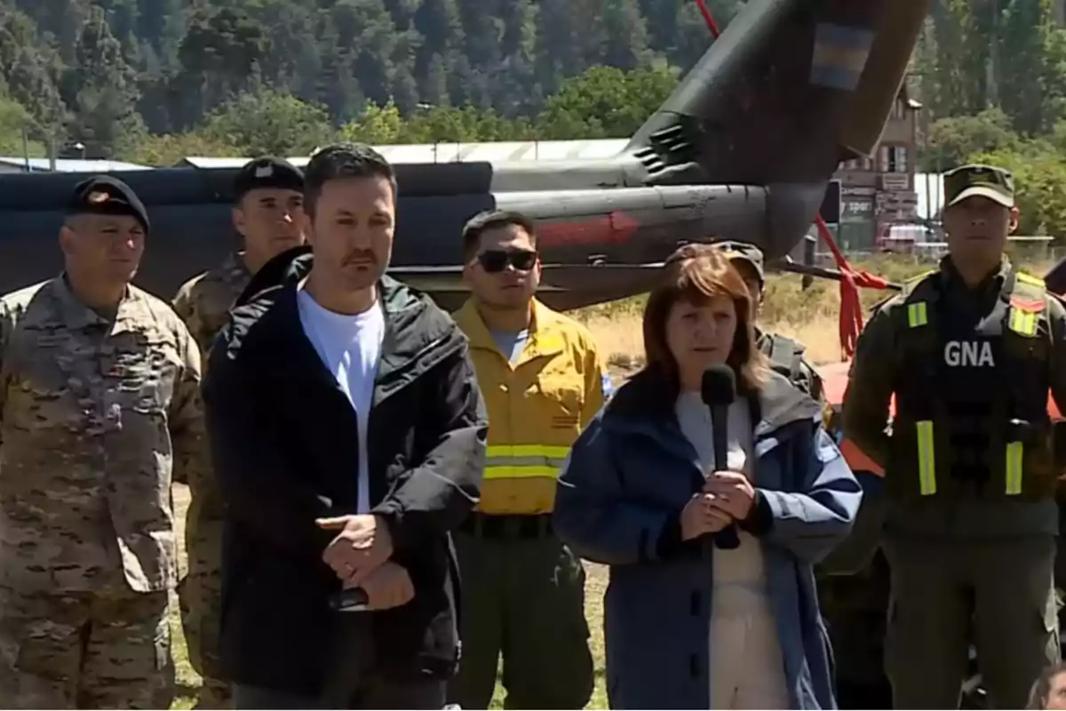 Un grupo de personas, algunas en uniforme militar, están de pie al aire libre frente a un helicóptero, con árboles al fondo.