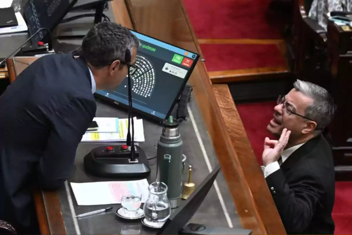 Dos personas conversan en un entorno de trabajo con pantallas de computadora y documentos sobre la mesa.