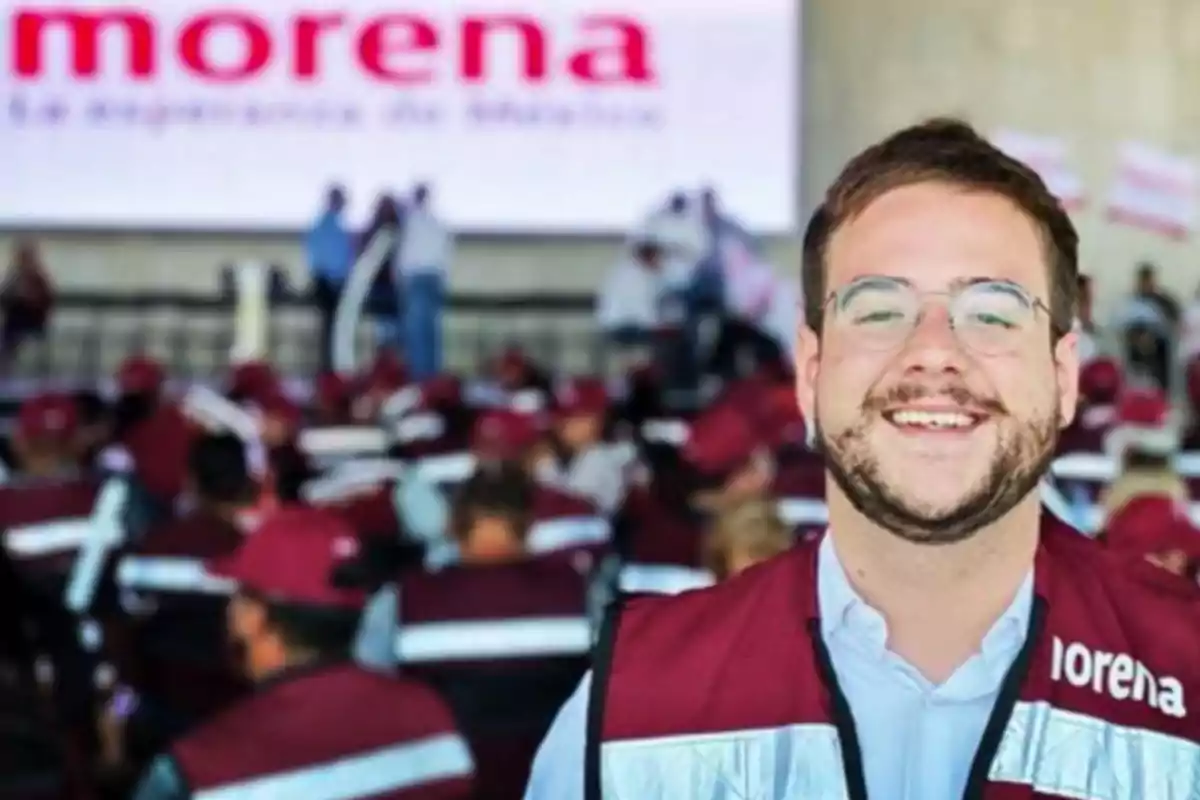 Un hombre sonriente con chaleco rojo y gafas en un evento de Morena con un grupo de personas al fondo.