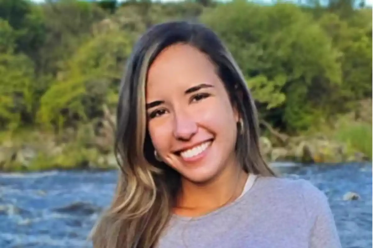 Una persona sonriendo al aire libre con un río y árboles de fondo.