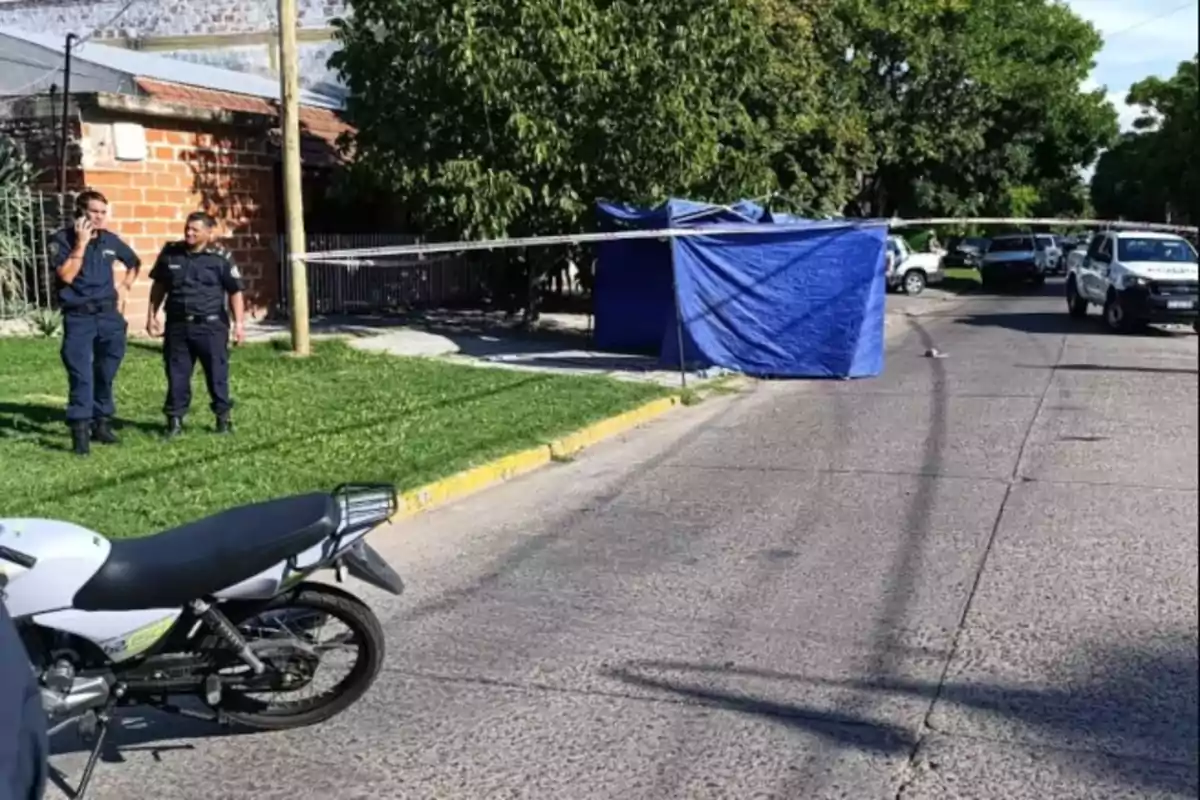 Una escena de calle con policías, una motocicleta y una carpa azul en un área acordonada.