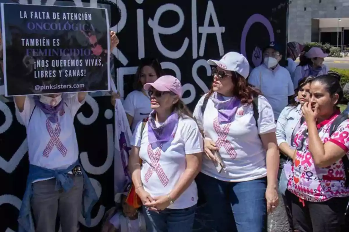 Un grupo de personas con camisetas blancas y pañuelos morados sostiene un cartel que destaca la falta de atención oncológica como un problema grave.