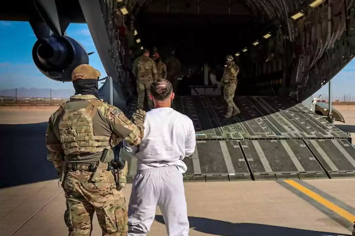 A group of military personnel escorts a man into a cargo plane on a runway.