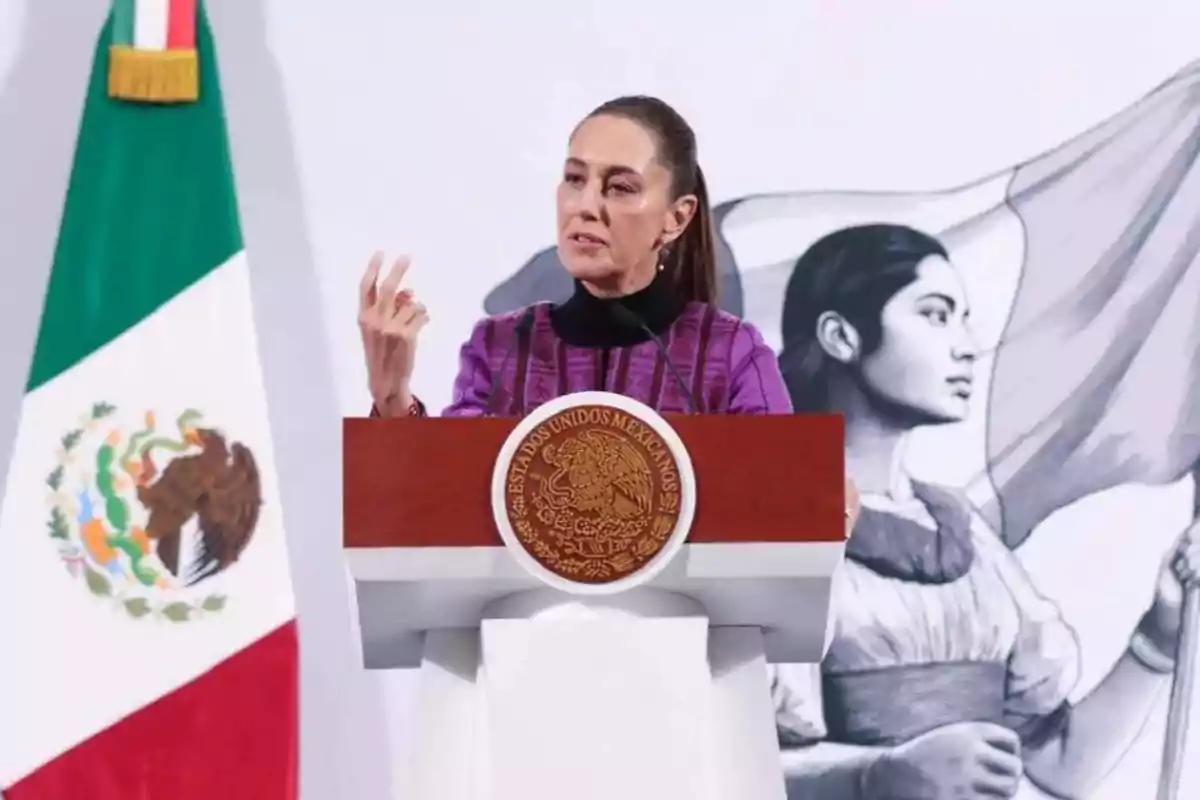 Una persona hablando en un podio con el escudo de México, una bandera mexicana a la izquierda y un mural de una mujer con una bandera al fondo.