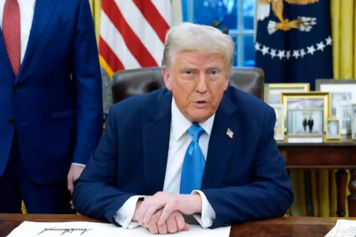 A man in a blue suit is sitting at a desk with a flag in the background.