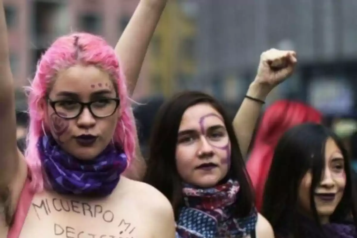 Un grupo de mujeres participa en una manifestación feminista, con una de ellas levantando el puño y mostrando un mensaje escrito en su cuerpo.