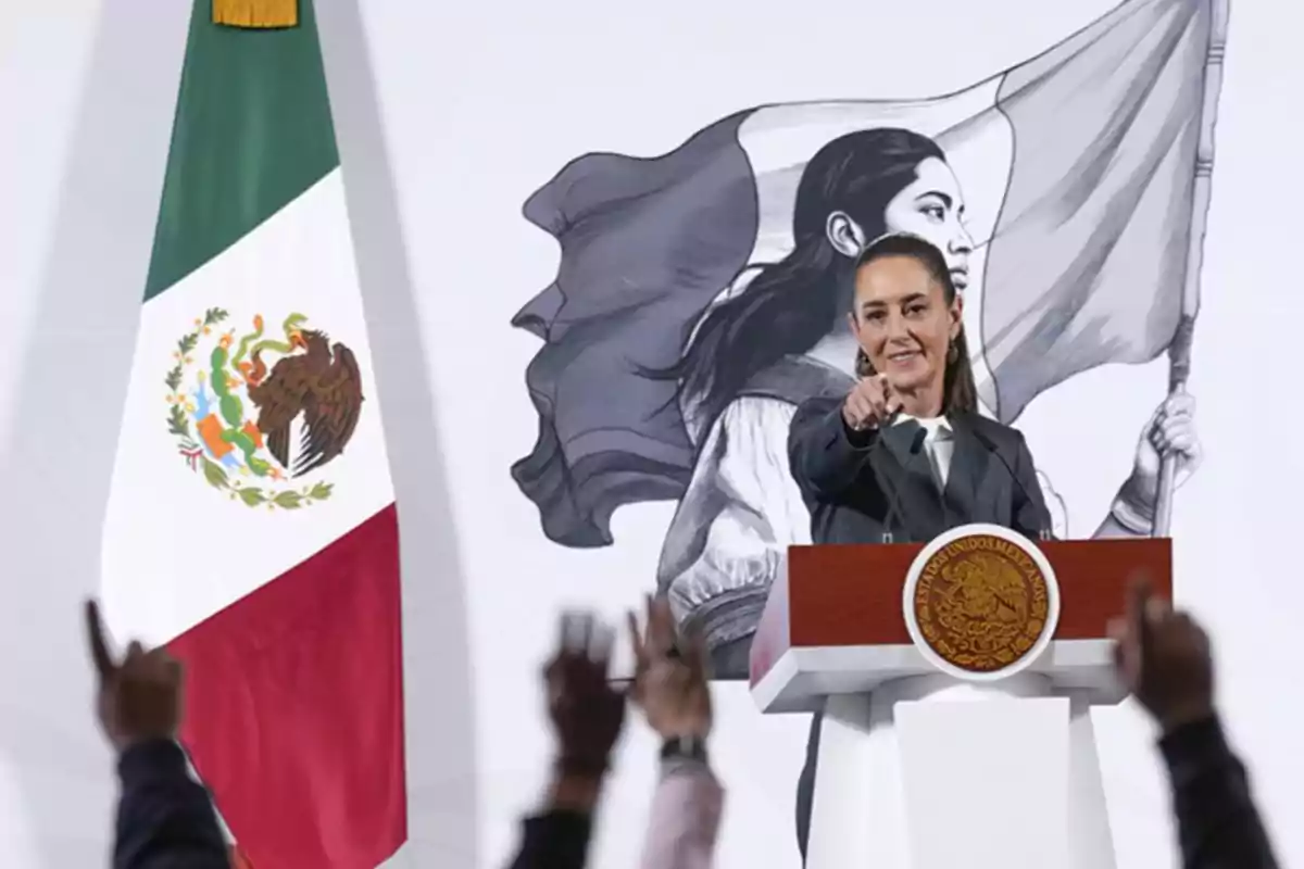 Una persona en un podio señalando hacia adelante, con una bandera de México a la izquierda y un mural de una mujer con una bandera al fondo.