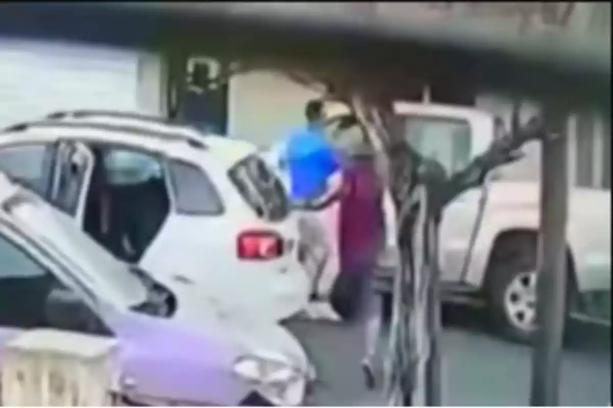 A person standing next to a white car with the rear door open on a street, while another vehicle is parked nearby.