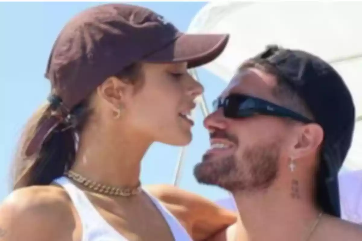 Dos personas con gorras y gafas de sol están sonriendo y mirándose de cerca en un ambiente al aire libre.