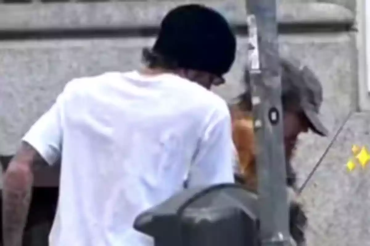 Two people standing on a sidewalk, one with a white t-jersey and black cap, the other with a cap, next to a metal post.