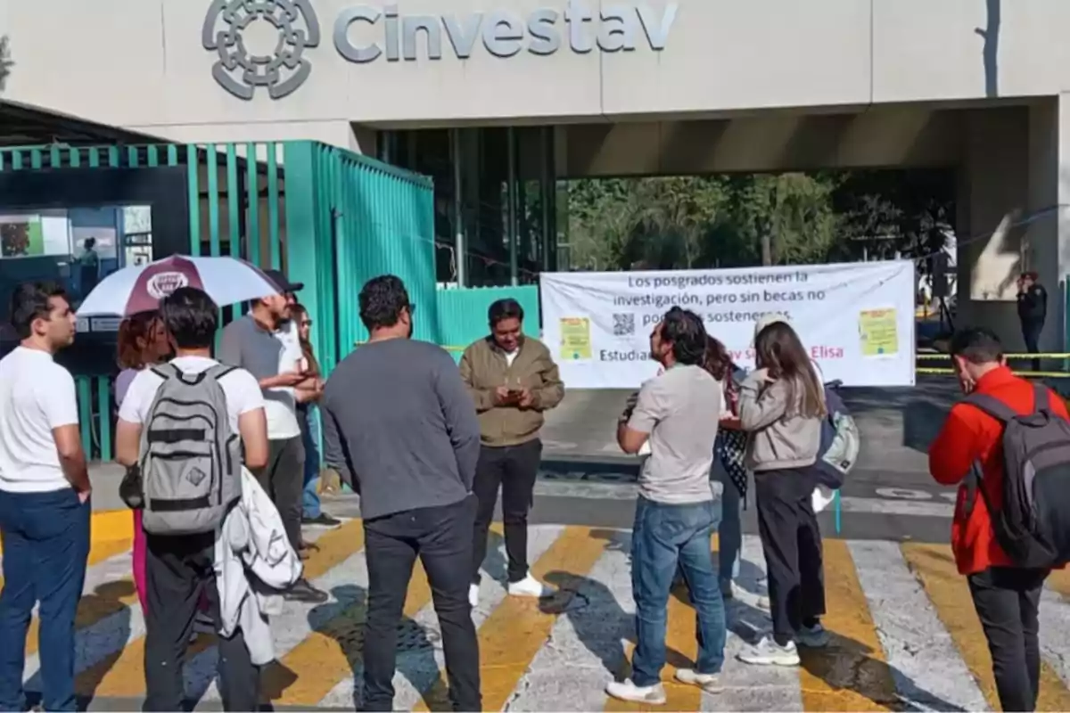 A group of people is gathered in front of the entrance of a building with the sign "Cinvestav," where there is a banner with a message about the importance of scholarships for graduate programs.