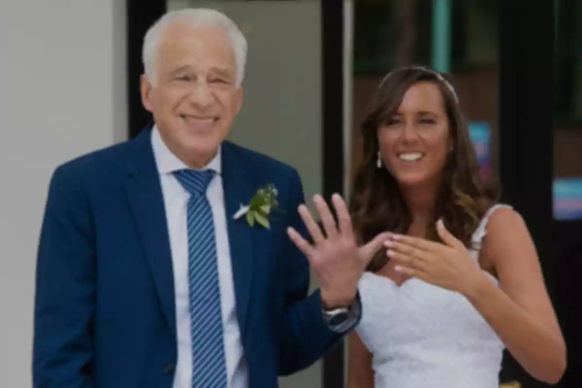 Un hombre mayor sonriente con traje azul y corbata a rayas junto a una mujer joven con vestido blanco, ambos haciendo un gesto con las manos.