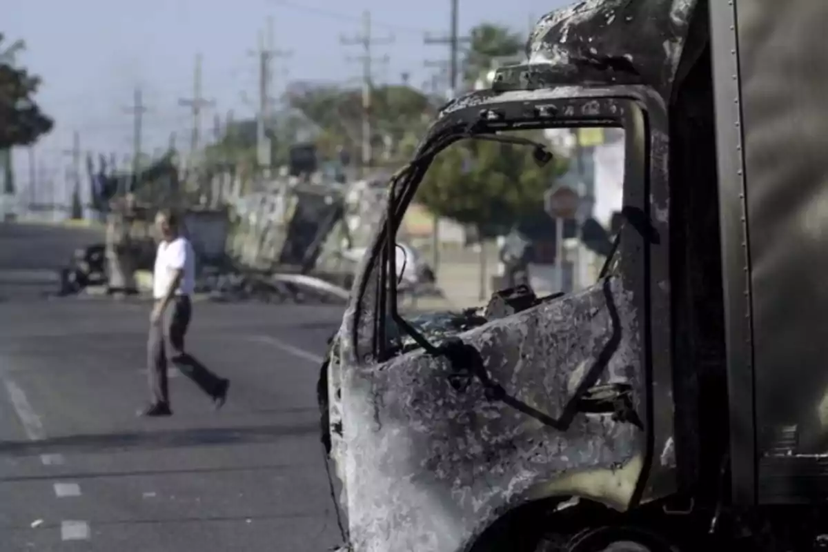 A burned truck on a street with a man walking in the background and other damaged vehicles in the surroundings.