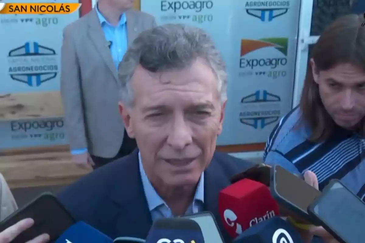 Un hombre hablando con varios micrófonos frente a él en un evento de Expoagro en San Nicolás.