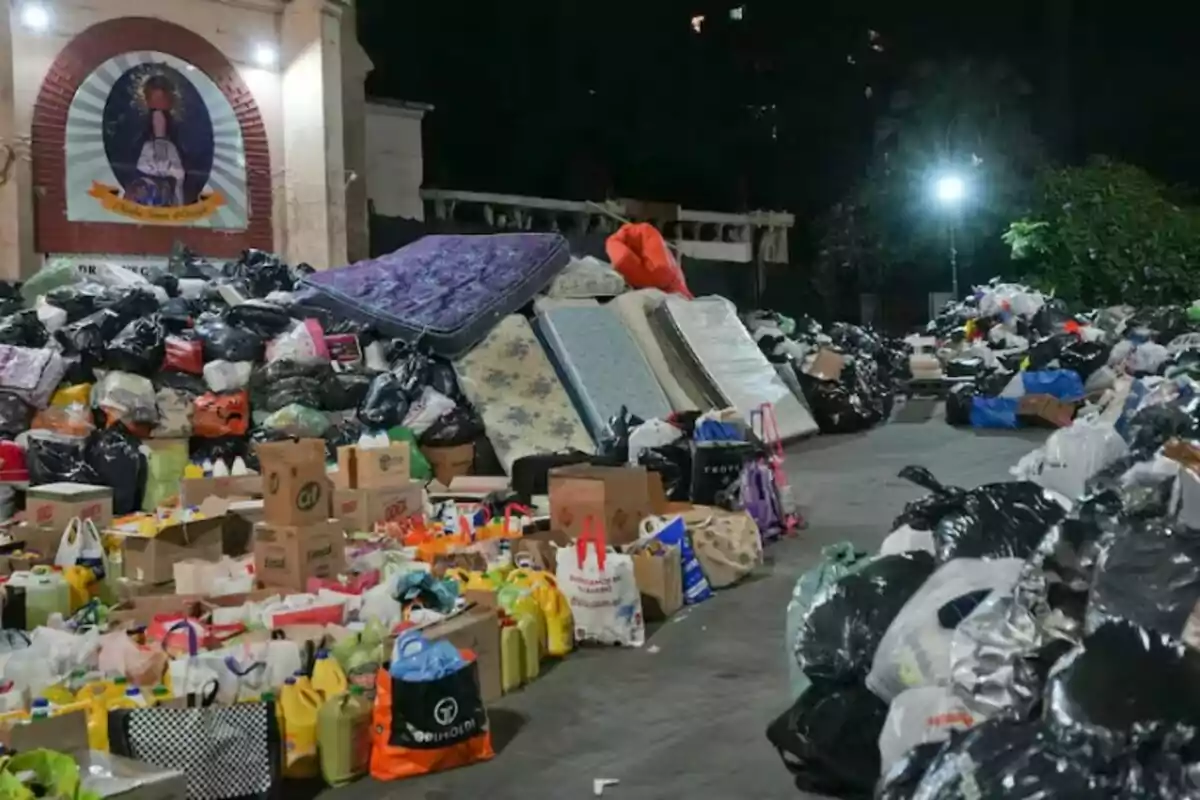 Una gran cantidad de bolsas de basura, colchones y cajas se acumulan en una plaza iluminada por la noche, con un mural religioso visible al fondo.