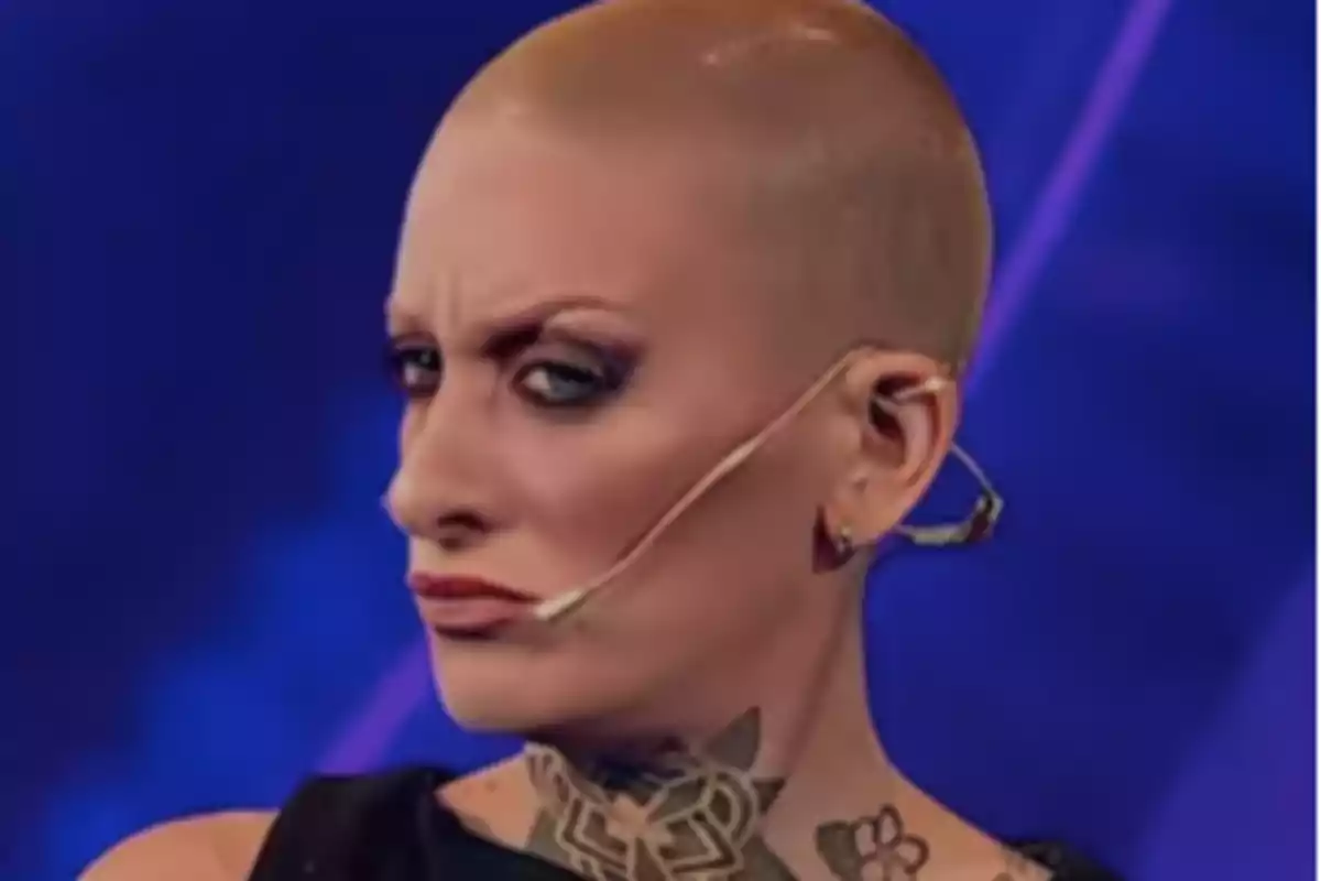 A person with a shaved head and dark makeup on a stage with a blue background.