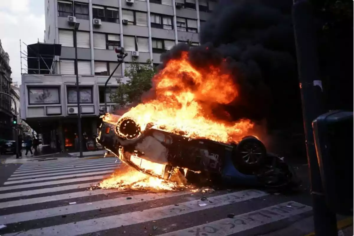 Un coche volcado en llamas en una intersección urbana con un edificio de fondo.