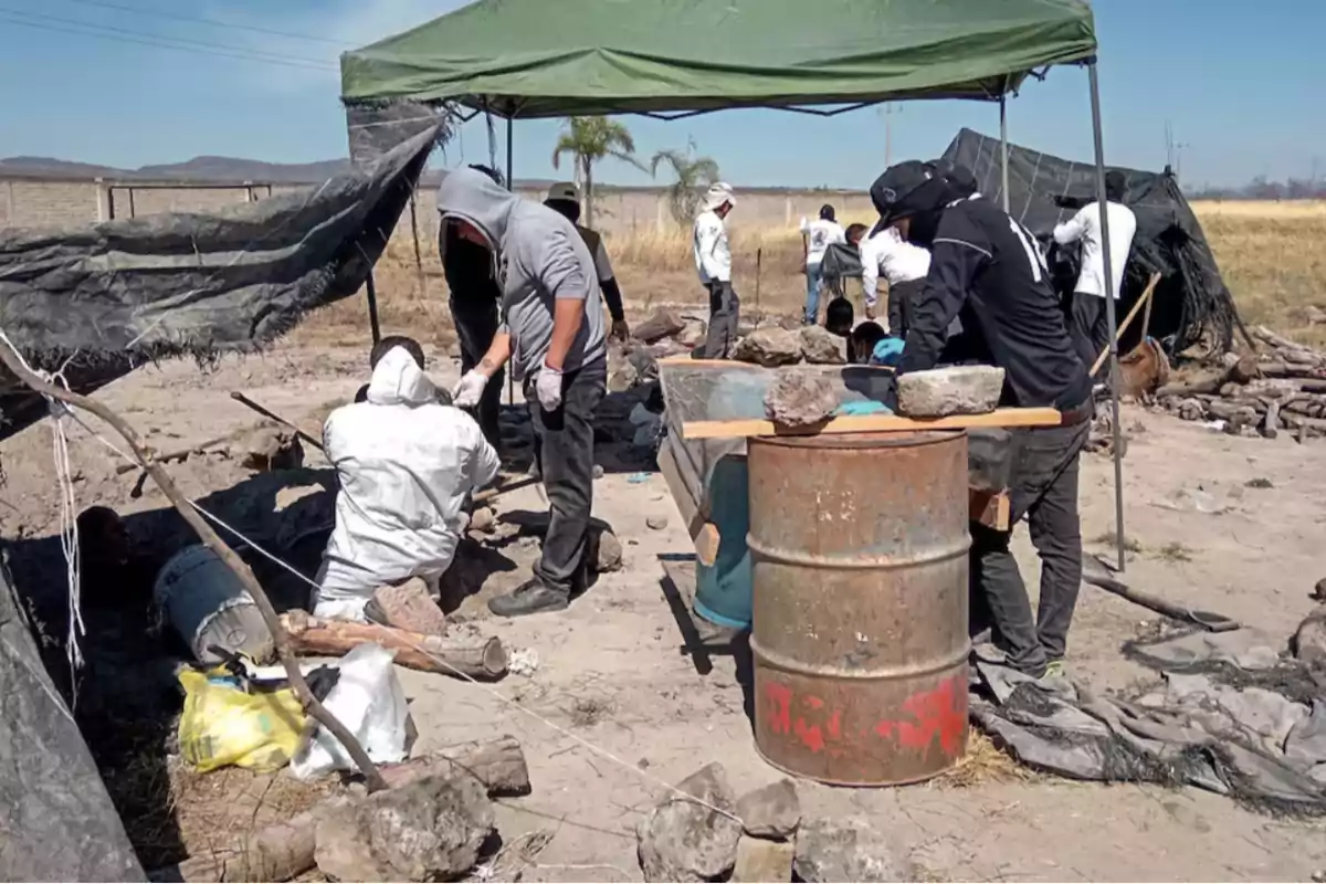 A group of people is working at an outdoor site under a green tent, surrounded by tools and construction materials.