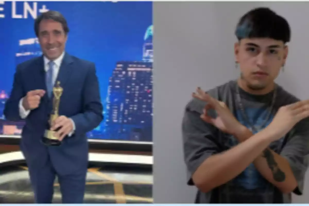 Un hombre con traje sostiene un trofeo en un estudio de televisión mientras otra persona hace un gesto con las manos frente a una pared blanca.