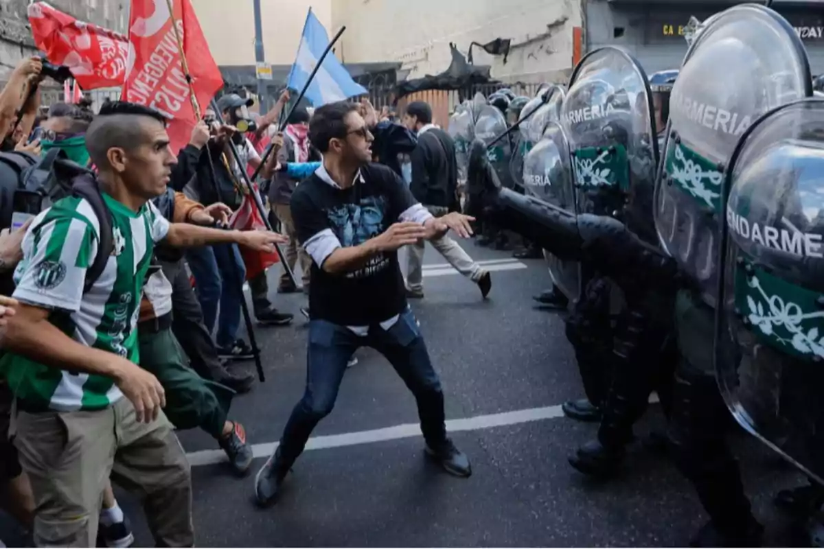 Un grupo de manifestantes se enfrenta a una línea de policías antidisturbios con escudos en una calle, mientras algunos sostienen banderas y otros intentan dialogar.