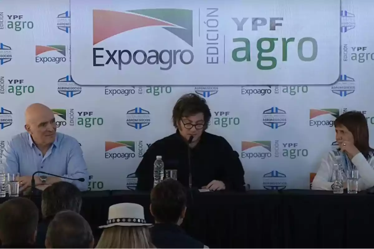 Tres personas sentadas en una mesa durante un evento de Expoagro con un fondo que muestra logotipos de YPF Agro y Expoagro.