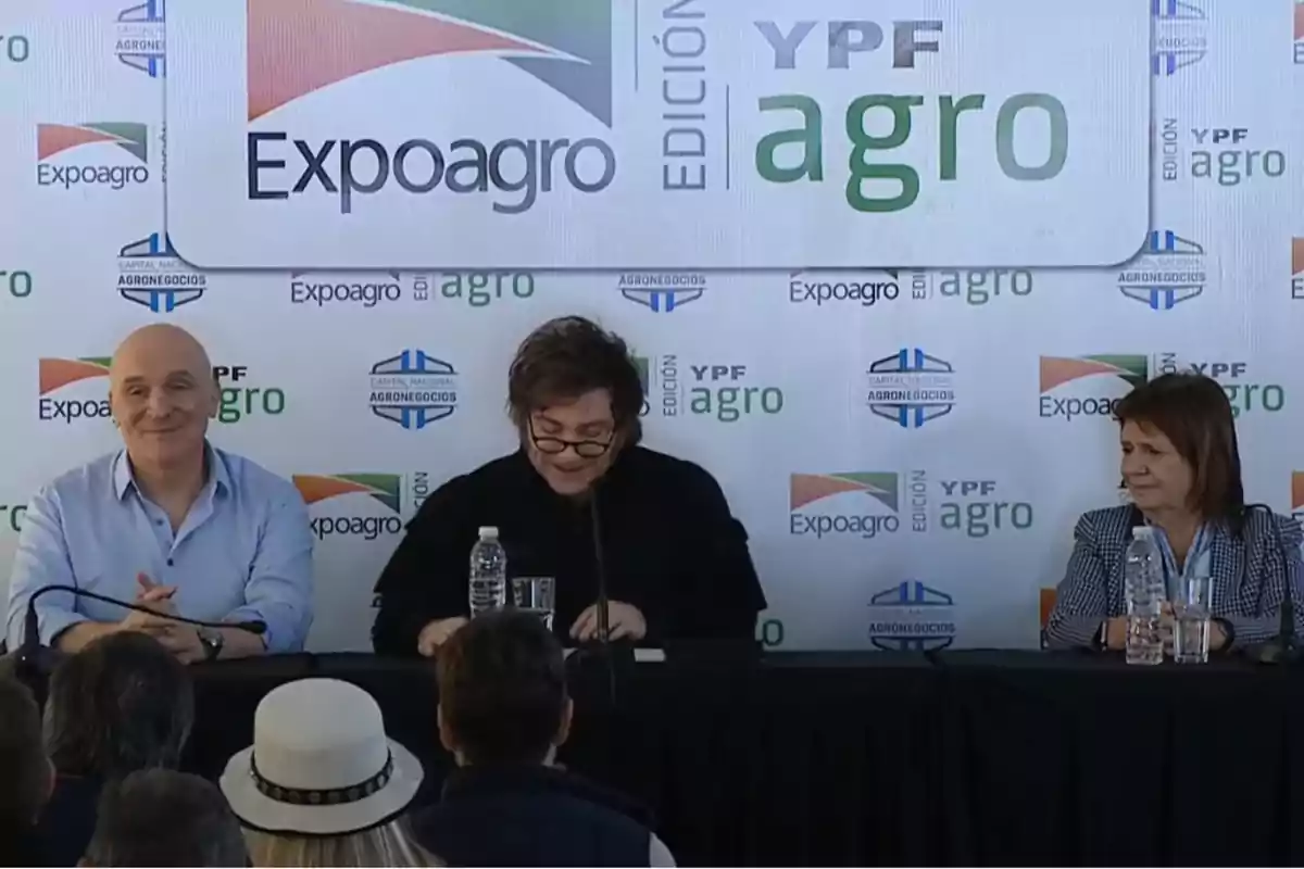 Tres personas están sentadas en una mesa durante un evento de Expoagro, con un fondo que muestra logotipos de Expoagro y YPF Agro.