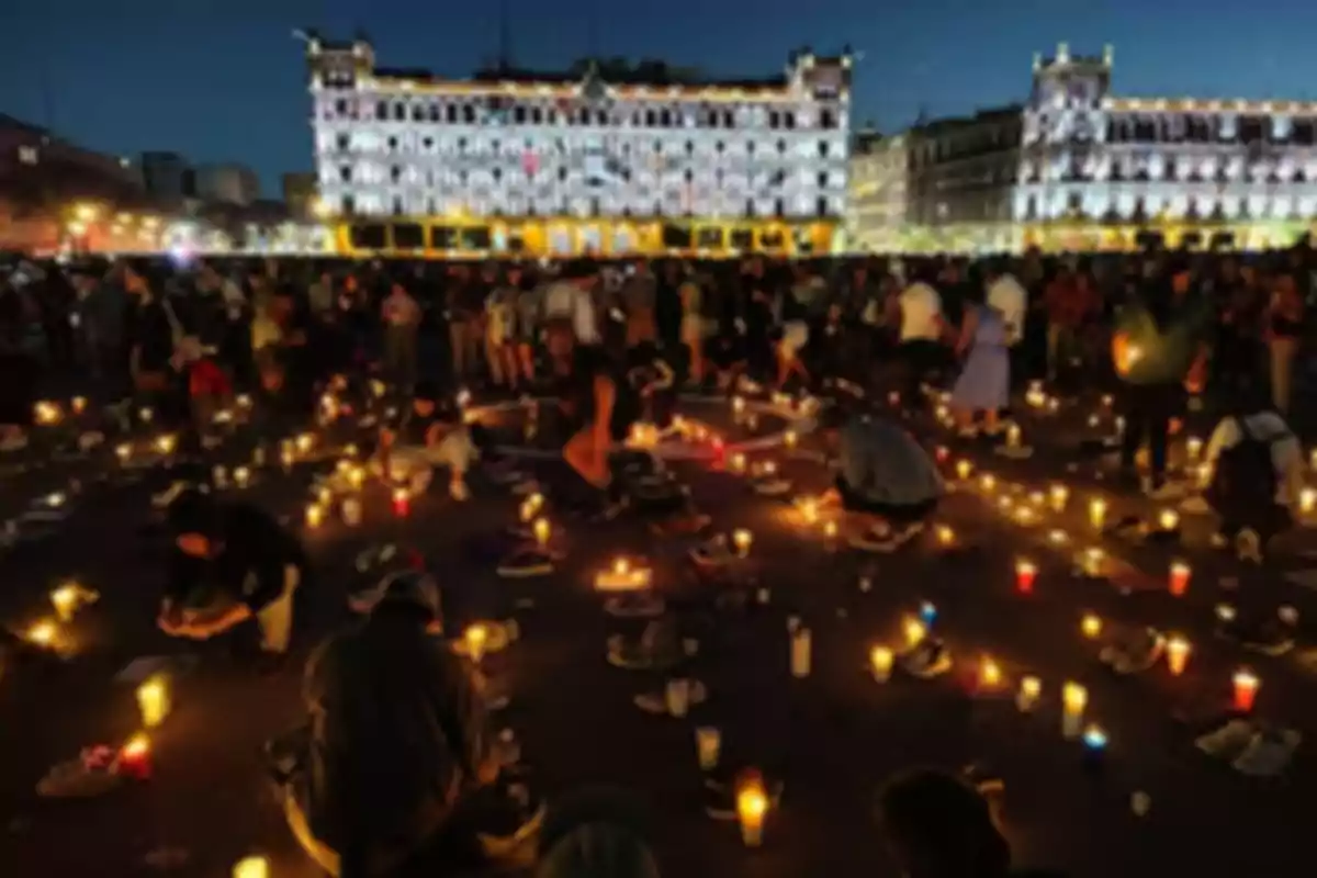 A crowd gathers in a candlelit square at night, with a historic building in the background.