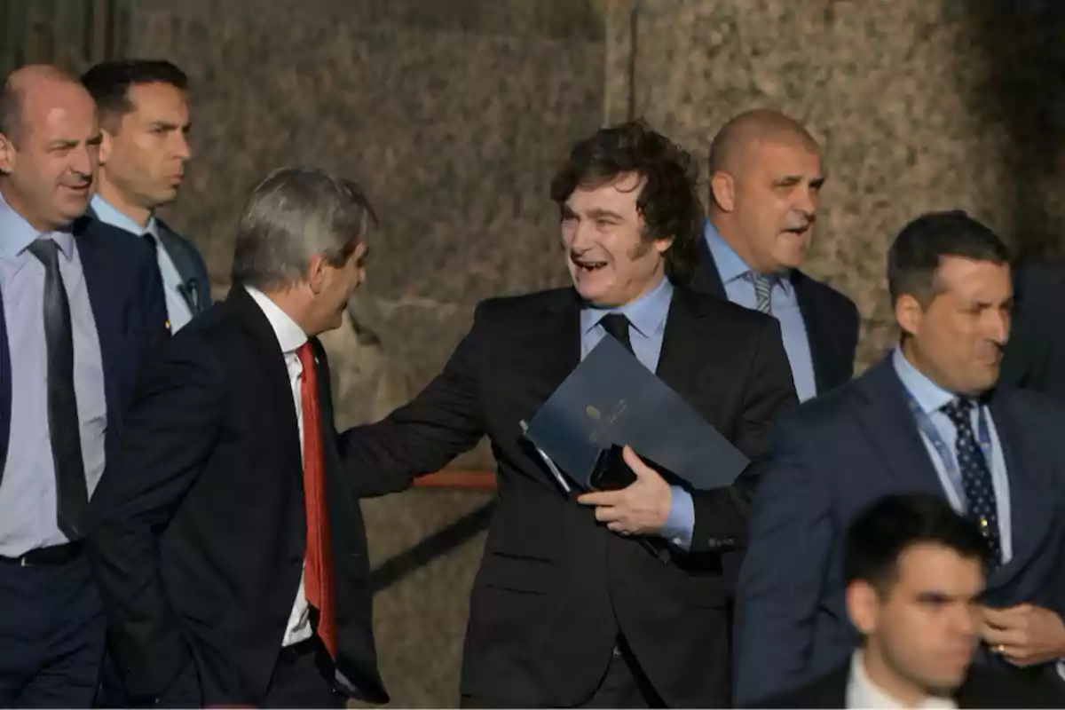 A group of men in formal suits walks outdoors while one of them smiles and holds a blue folder. A group of men in formal suits walks outdoors while one of them smiles and holds a blue folder.
