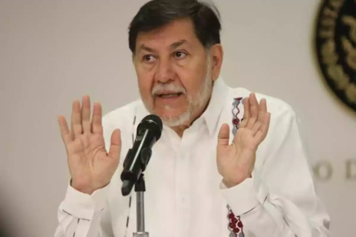 Un hombre con barba y camisa blanca habla frente a un micrófono con las manos levantadas.