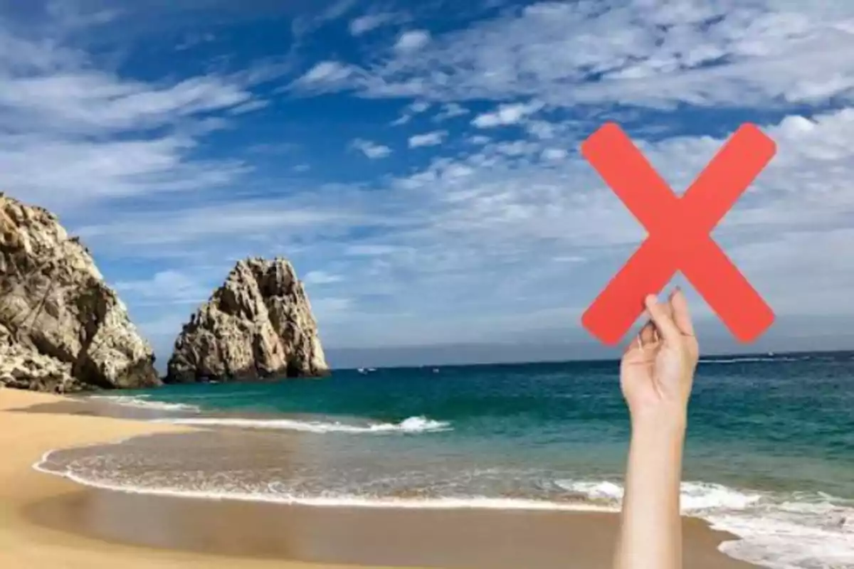 Una mano sostiene una gran equis roja frente a un paisaje de playa con formaciones rocosas y cielo azul.