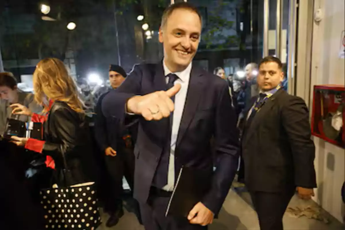 A smiling man in a dark suit gives a thumbs-up gesture while walking through a crowded place.
