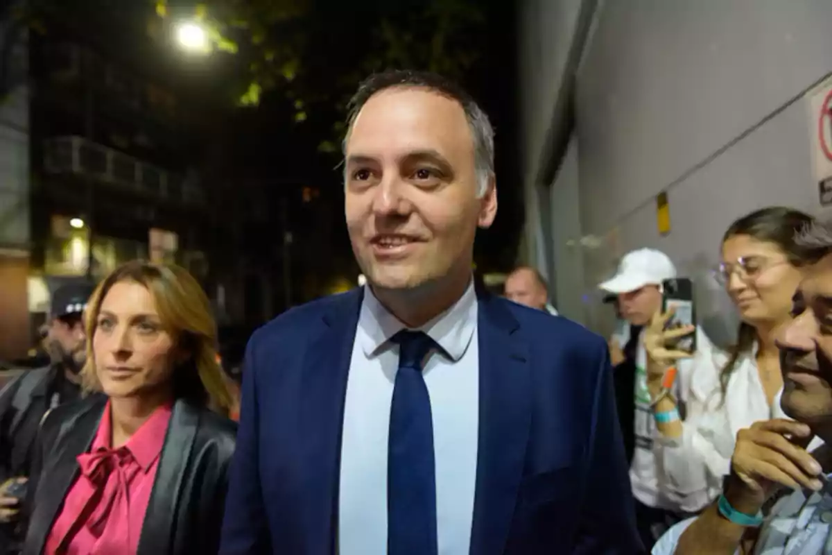 A man in a blue suit and tie walks down the street surrounded by people, some of whom are taking pictures with their phones. A man in a blue suit and tie walks down the street surrounded by people, some of whom are taking pictures with their phones.