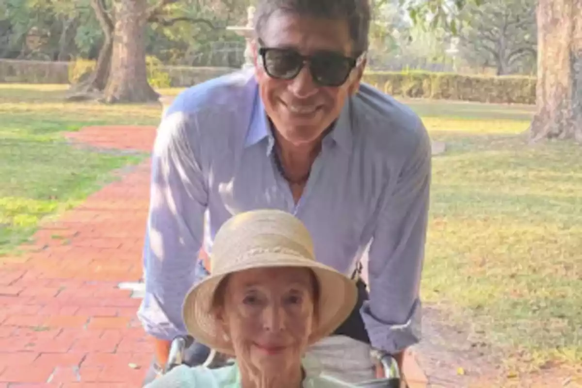 A man with sunglasses pushes an elderly woman in a wheelchair in a park.
